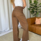 Woman wearing RFM Full Size High Rise Garment Dye Wide Leg Jeans in brown, standing by a brown leather couch and green plant.