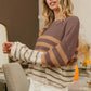 Woman wearing a trendy BiBi color block striped long sleeve sweater in earthy tones, paired with white shorts.