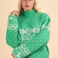 Woman wearing green Christmas sweater with snowflake and reindeer design, mock neck, and long sleeves, paired with a gray cap.