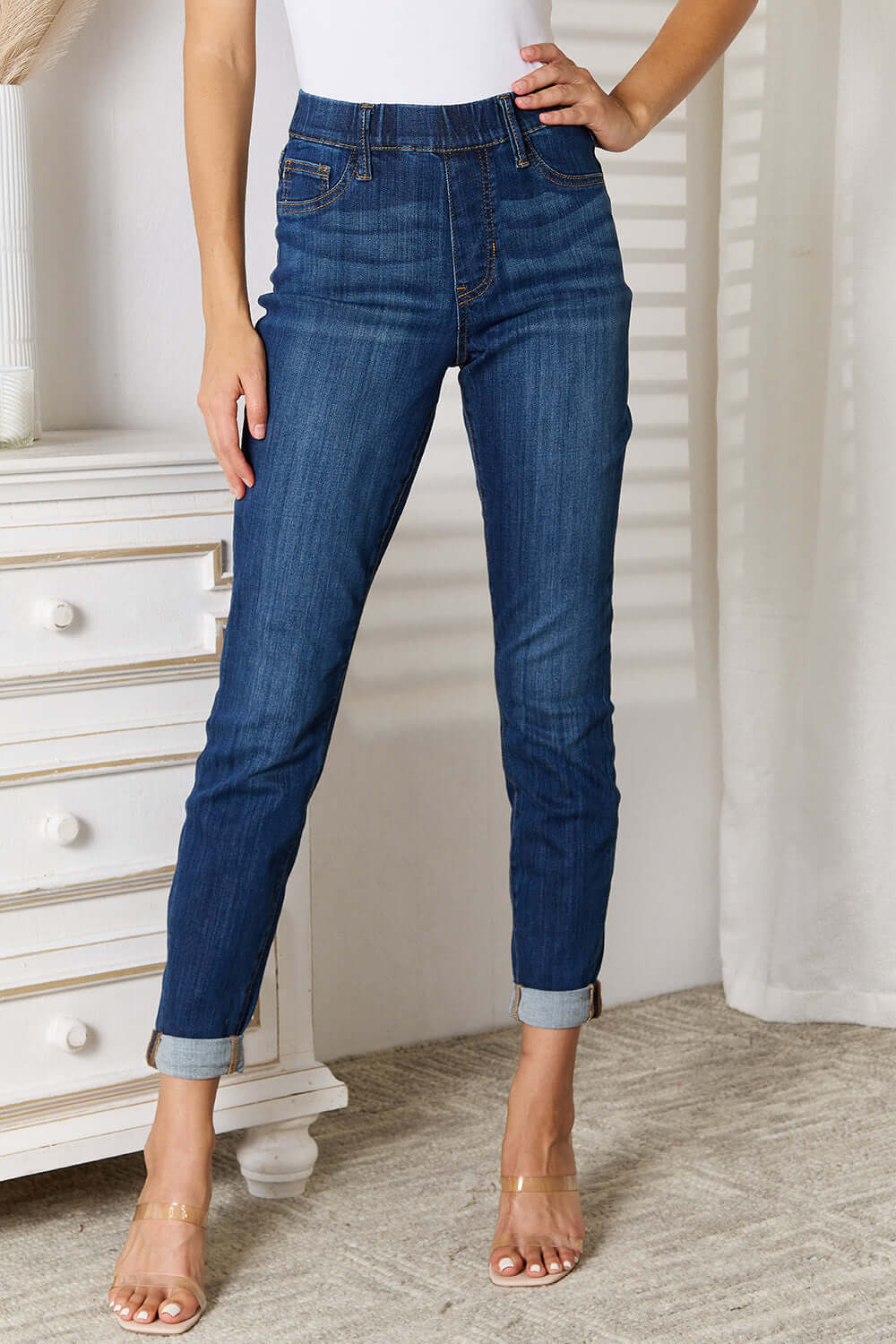 Woman wearing Judy Blue skinny cropped jeans with moderate stretch, paired with clear heels, standing in a stylish, modern room.