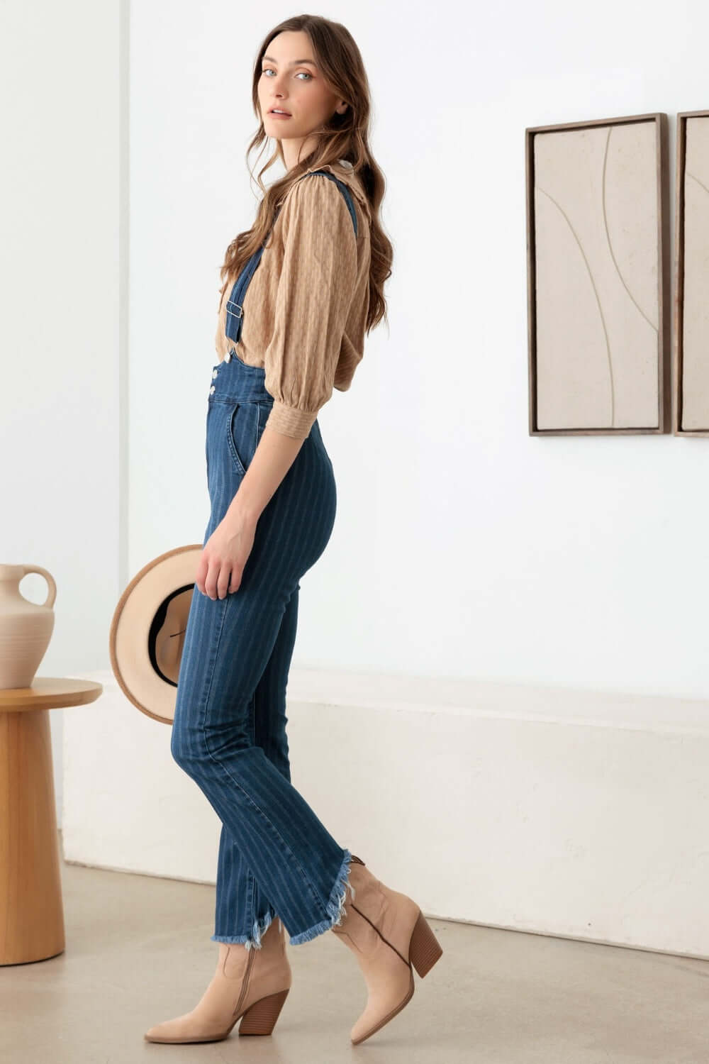 Woman wearing striped stretched suspender denim overalls with destroyed hem and high waist design, posing stylishly indoors.