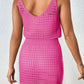 Woman wearing a pink openwork V-neck sleeveless cover up dress made of 100% acrylic, showing back view against white textured wall.