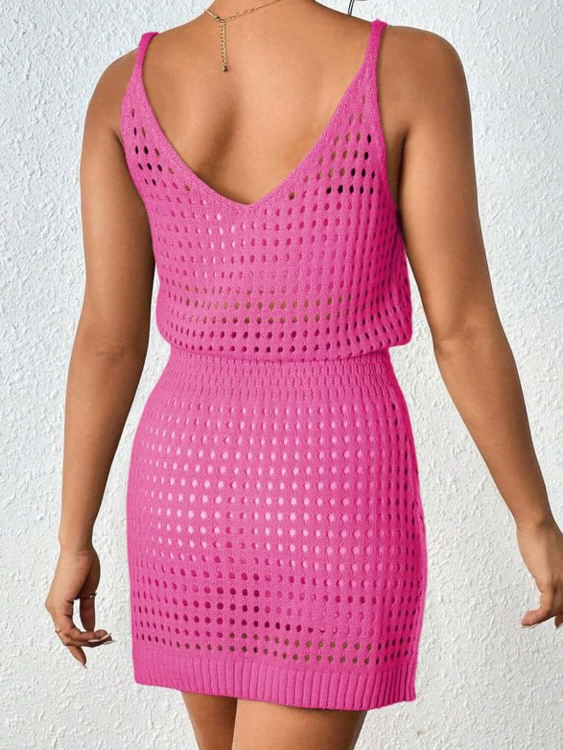 Woman wearing a pink openwork V-neck sleeveless cover up dress made of 100% acrylic, showing back view against white textured wall.