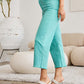 Woman wearing RFM Jeans Crop Chloe Full Size Tummy Control High Waist Raw Hem Jeans in teal, paired with white heels in a modern living room.