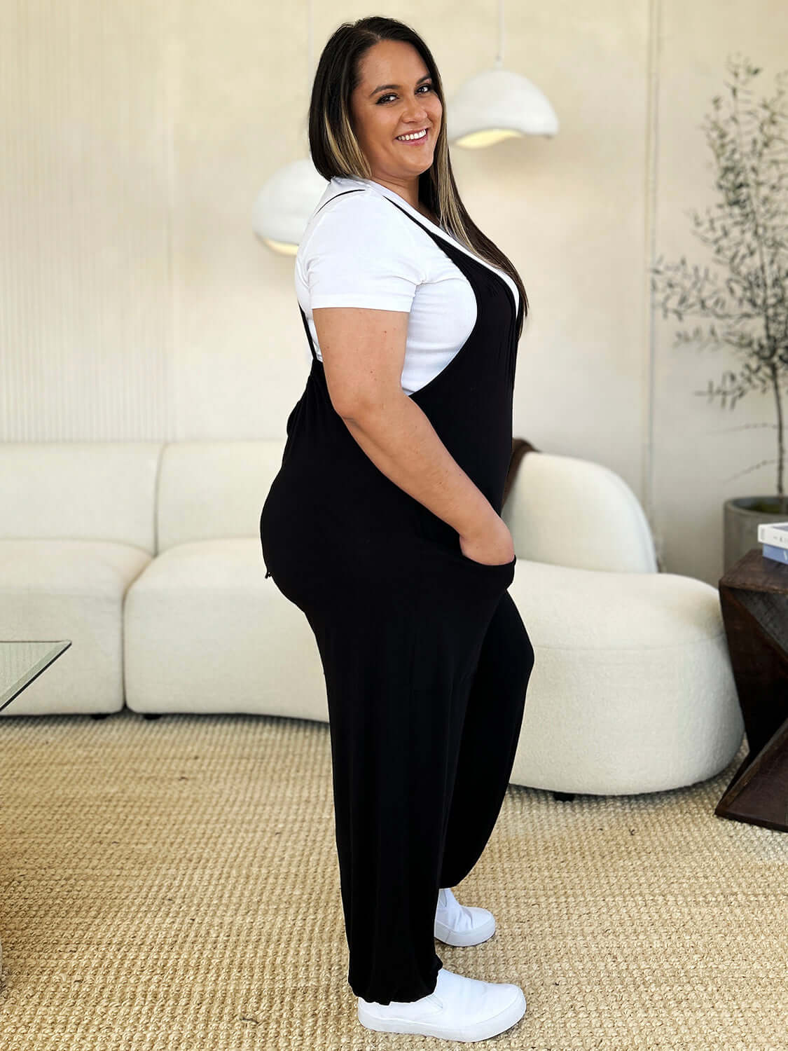 Woman wearing a Plunge Sleeveless Jumpsuit with Pockets, paired with a white shirt and white sneakers, standing in a modern living room.