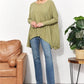 Woman wearing an oversized super soft rib layering top with a sharkbite hem and round neck, paired with jeans, standing by a plant and chair.