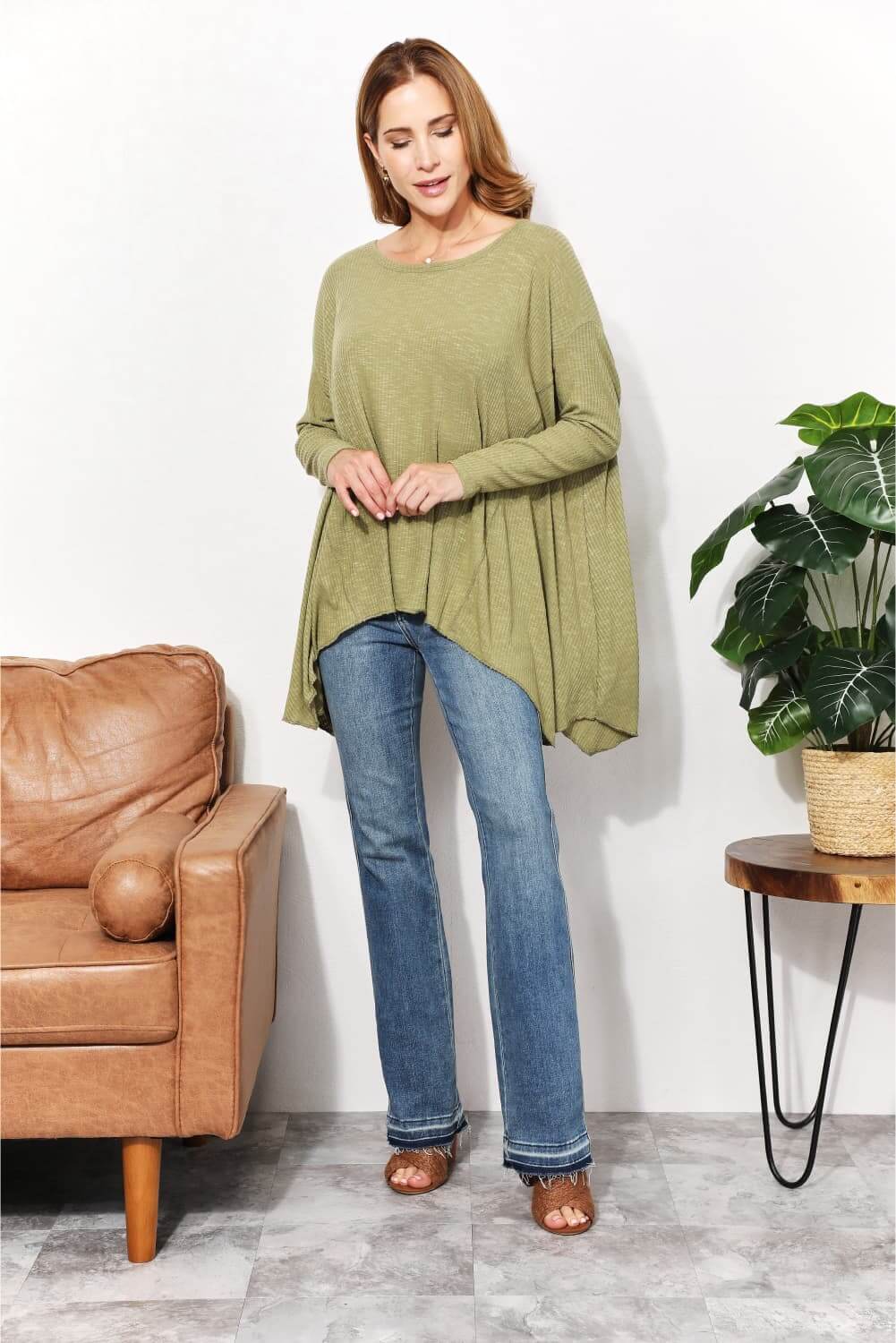 Woman wearing an oversized super soft rib layering top with a sharkbite hem and round neck, paired with jeans, standing by a plant and chair.