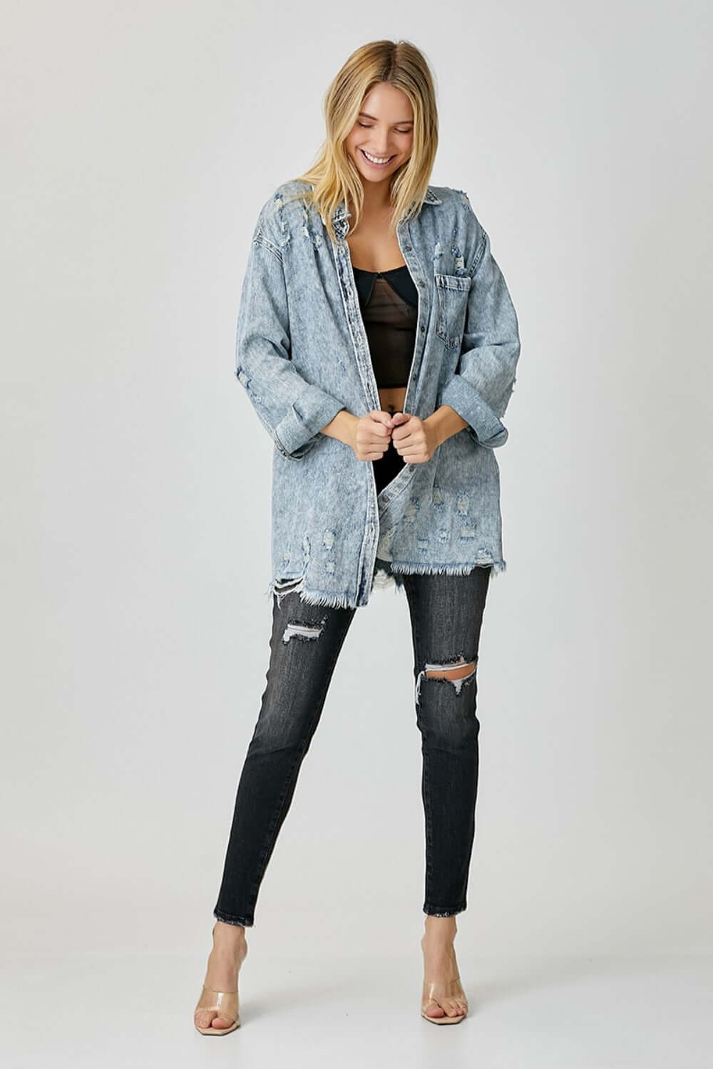 Woman wearing a distressed raw hem denim shirt by Risen Jeans, styled with black ripped jeans and nude heels, exuding casual and edgy vibes.