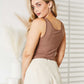 Woman wearing Everyday Full Size Basic Tank Bodysuit in brown, looking back, styled with white high-waisted shorts in a bright room.