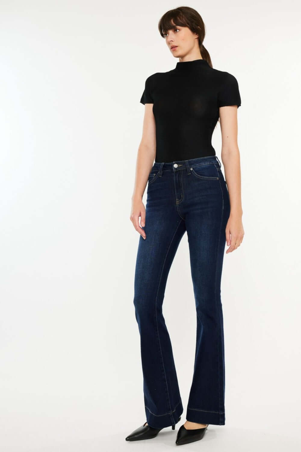 Woman wearing Kancan High Rise Slim Flare Jeans, dark stone wash denim with a sleek fit, paired with a black top and heels.