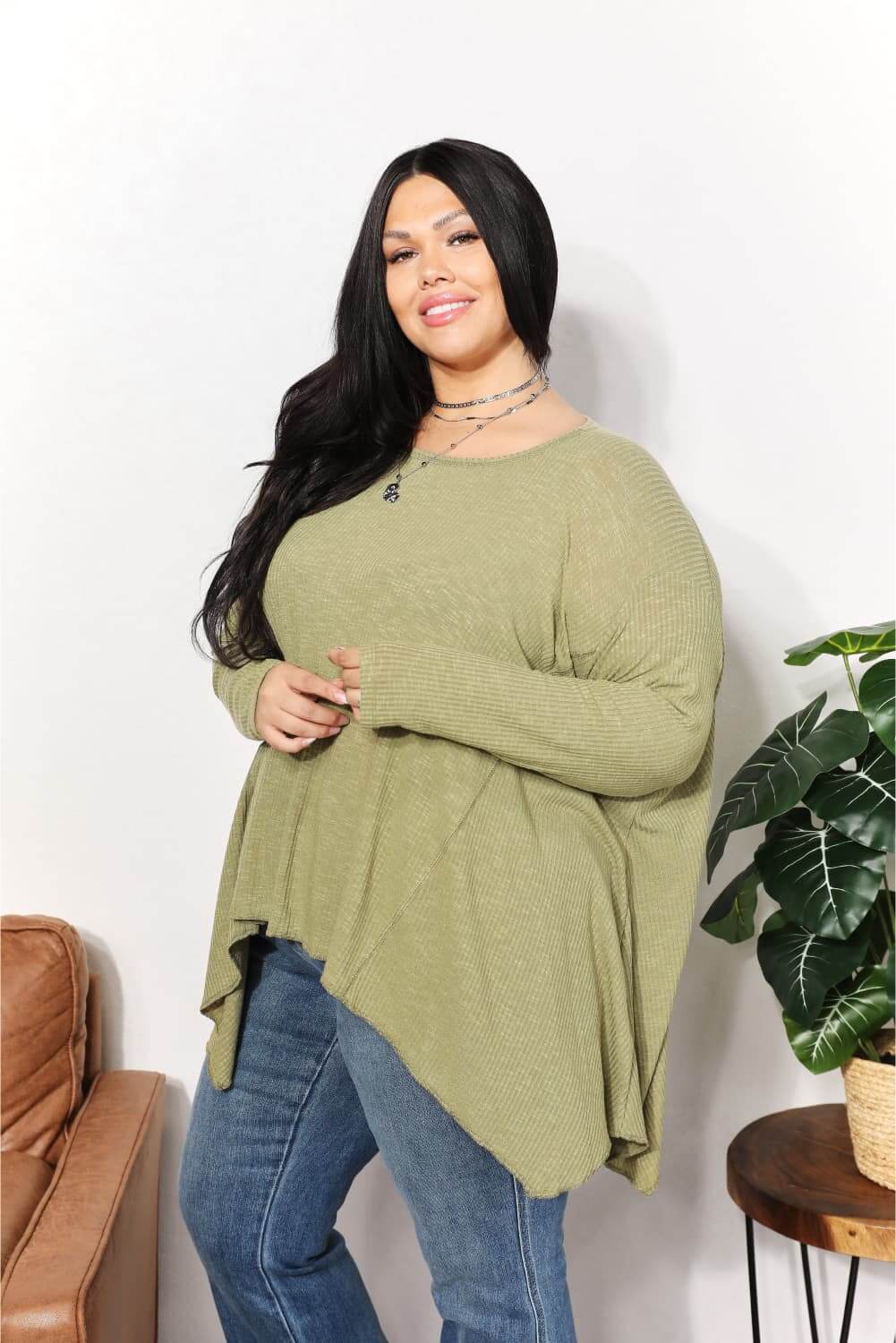 Woman wearing oversized super soft rib layering top with sharkbite hem and round neck, styled with jeans, standing next to a plant.