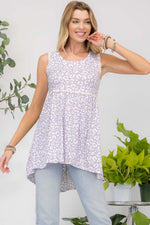 Woman wearing a Contrast High-Low Hem Tank with a floral pattern, paired with light-wash jeans, standing next to green potted plants.