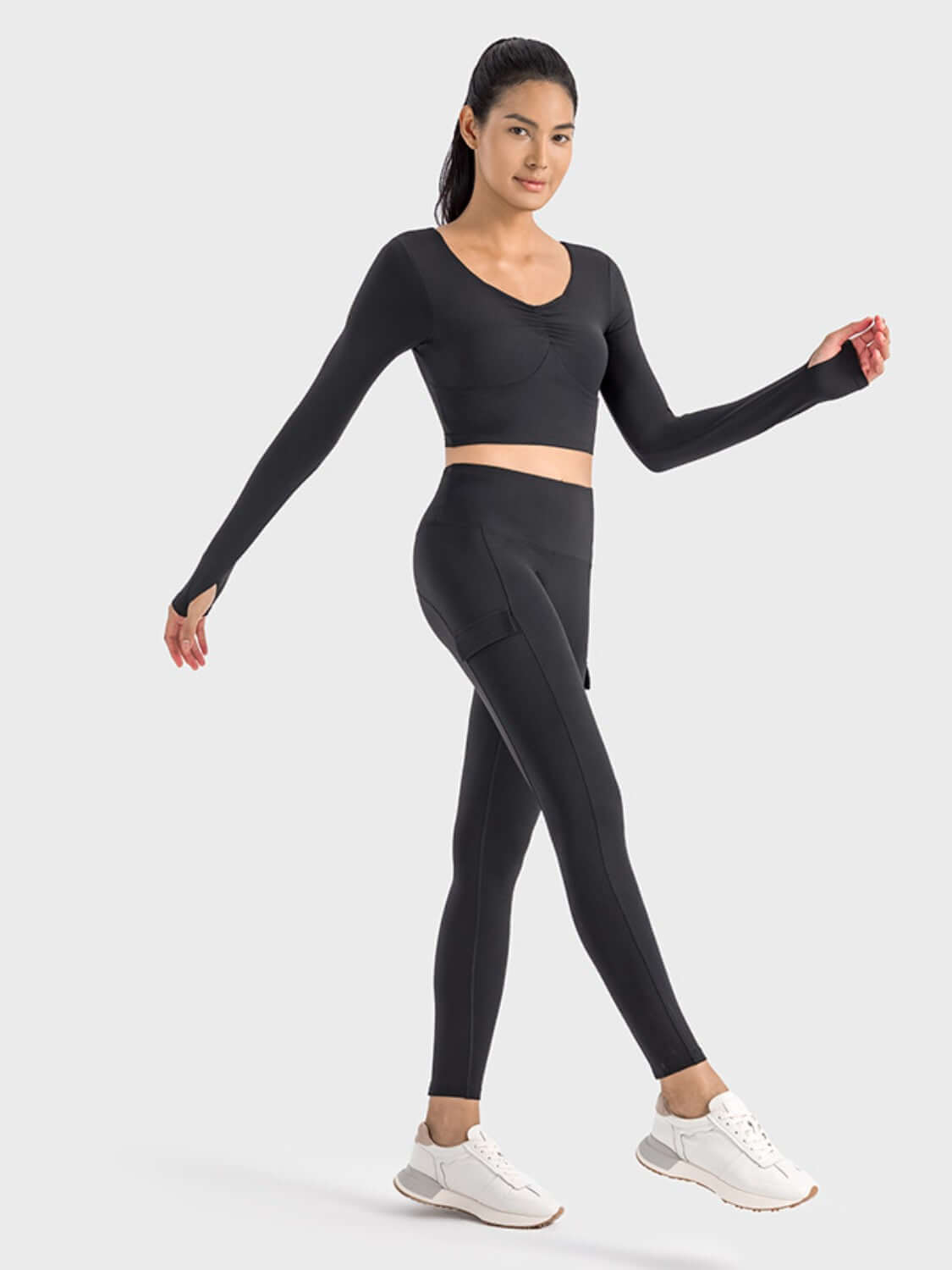 Model showcasing the Millennia Ruched Cropped Long Sleeve Sports Top in black, paired with matching leggings and sporty sneakers.