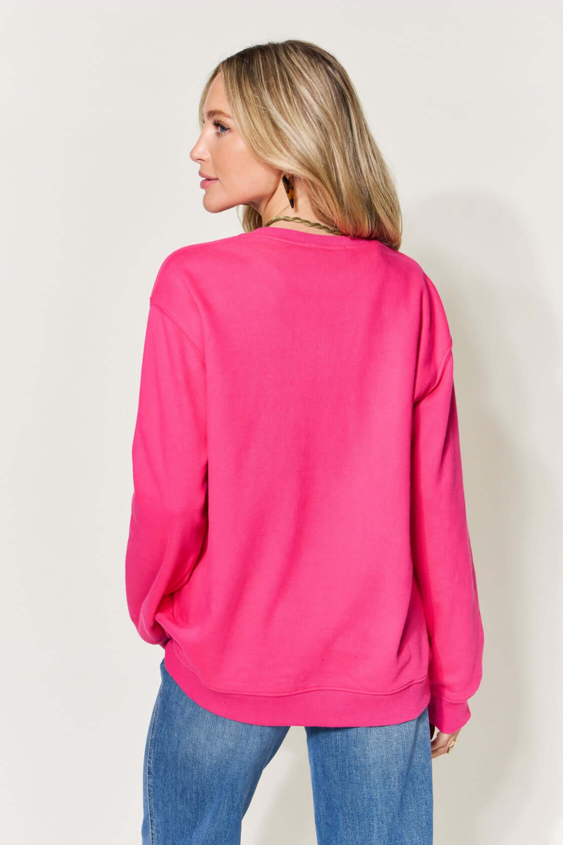 Woman wearing a pink long sleeve sweatshirt with letter graphic, paired with blue jeans, shown from the back.