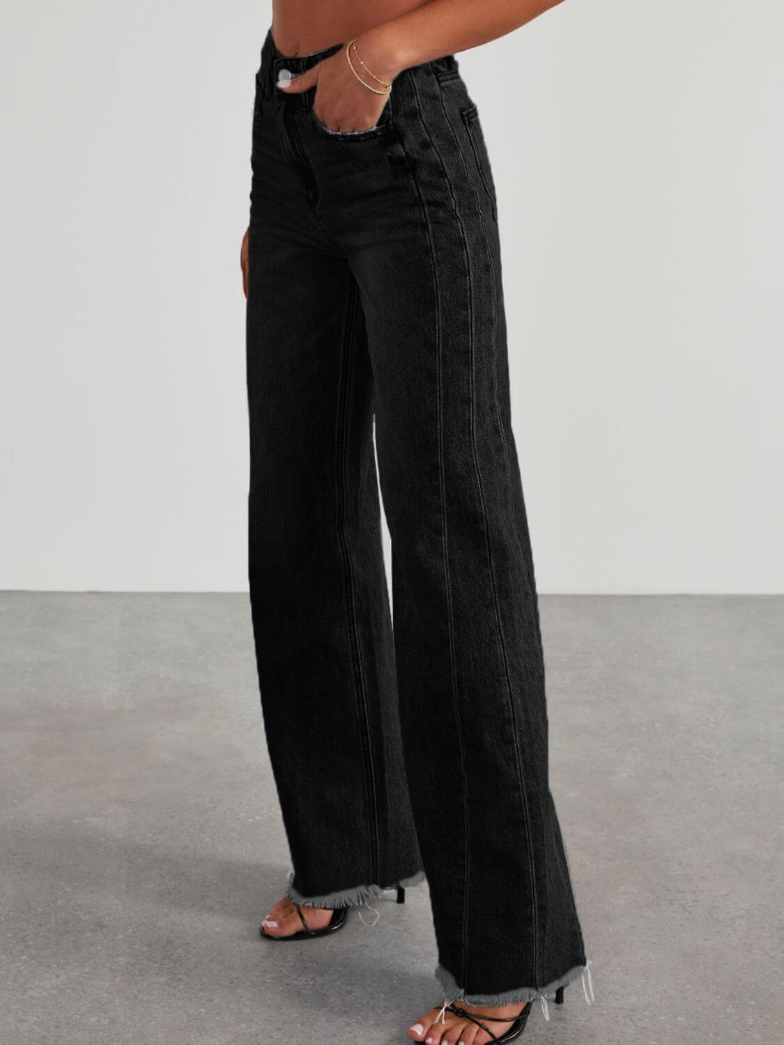 Woman wearing Bella Road Raw Hem Wide Leg Jeans with Pockets, black, non-stretch denim, side view, paired with heels.