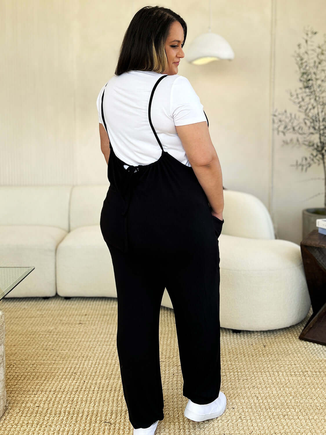 Woman wearing a black sleeveless jumpsuit with a white t-shirt, featuring a plunge neckline and convenient pockets, styled with casual shoes.