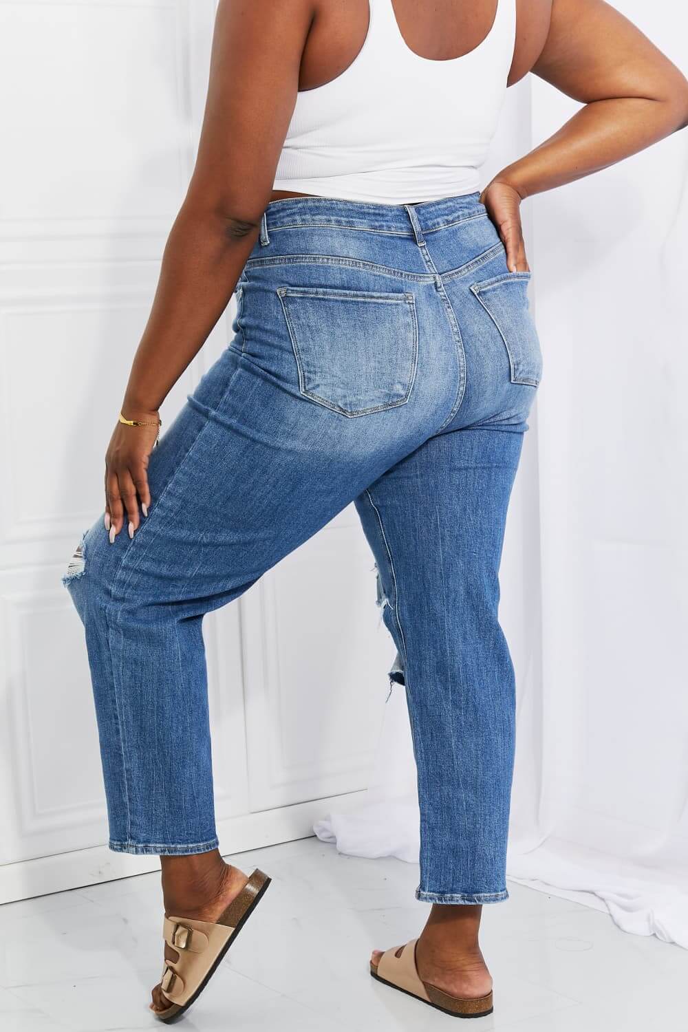 Back view of Emily High Rise Relaxed Jeans in blue with distressed knees, featuring a casual cropped fit, worn with a white top. Risen Jeans.