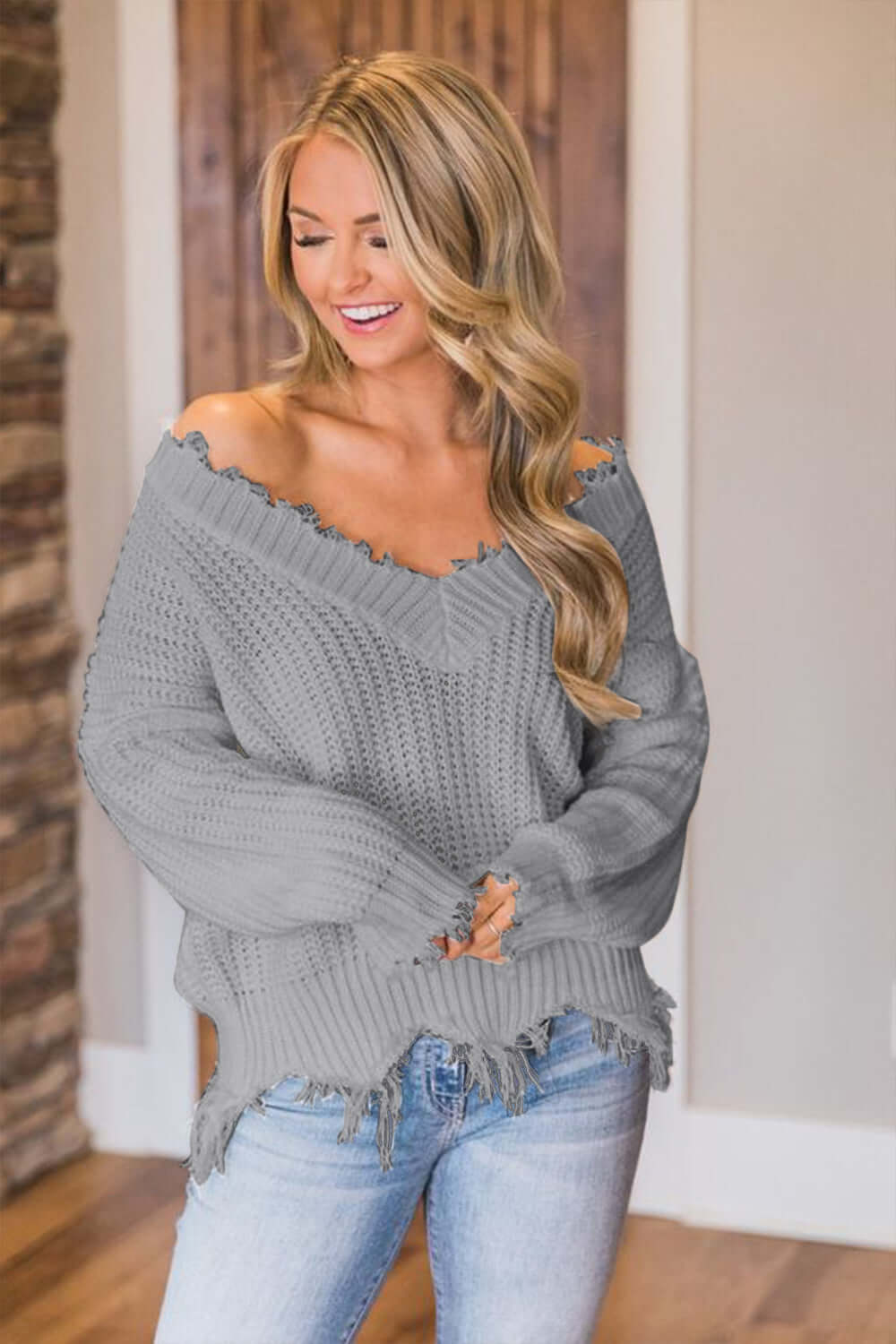 Woman wearing Bella Road frayed hem dropped shoulder sweater in grey, smiling indoors.