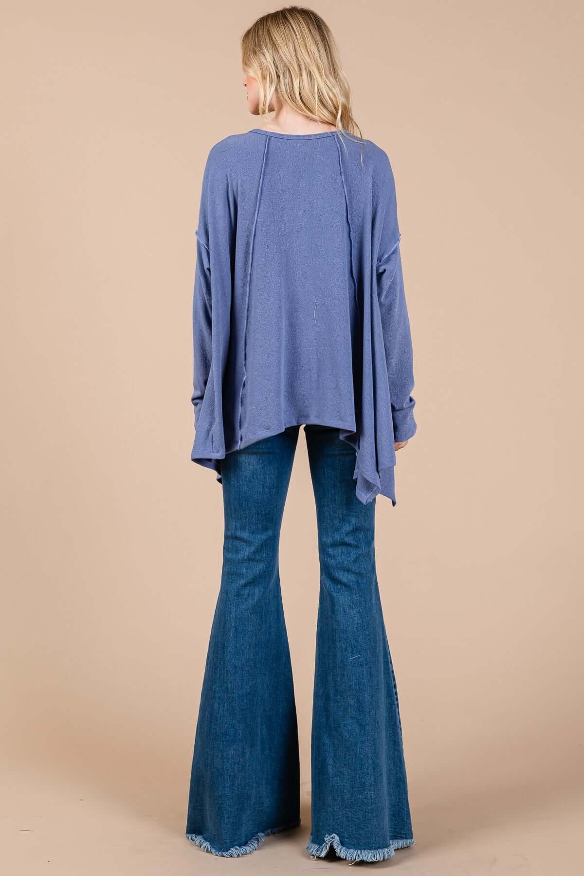 Woman wearing purple asymmetrical long sleeve top with drop sleeves, showing back view, paired with flared blue jeans.