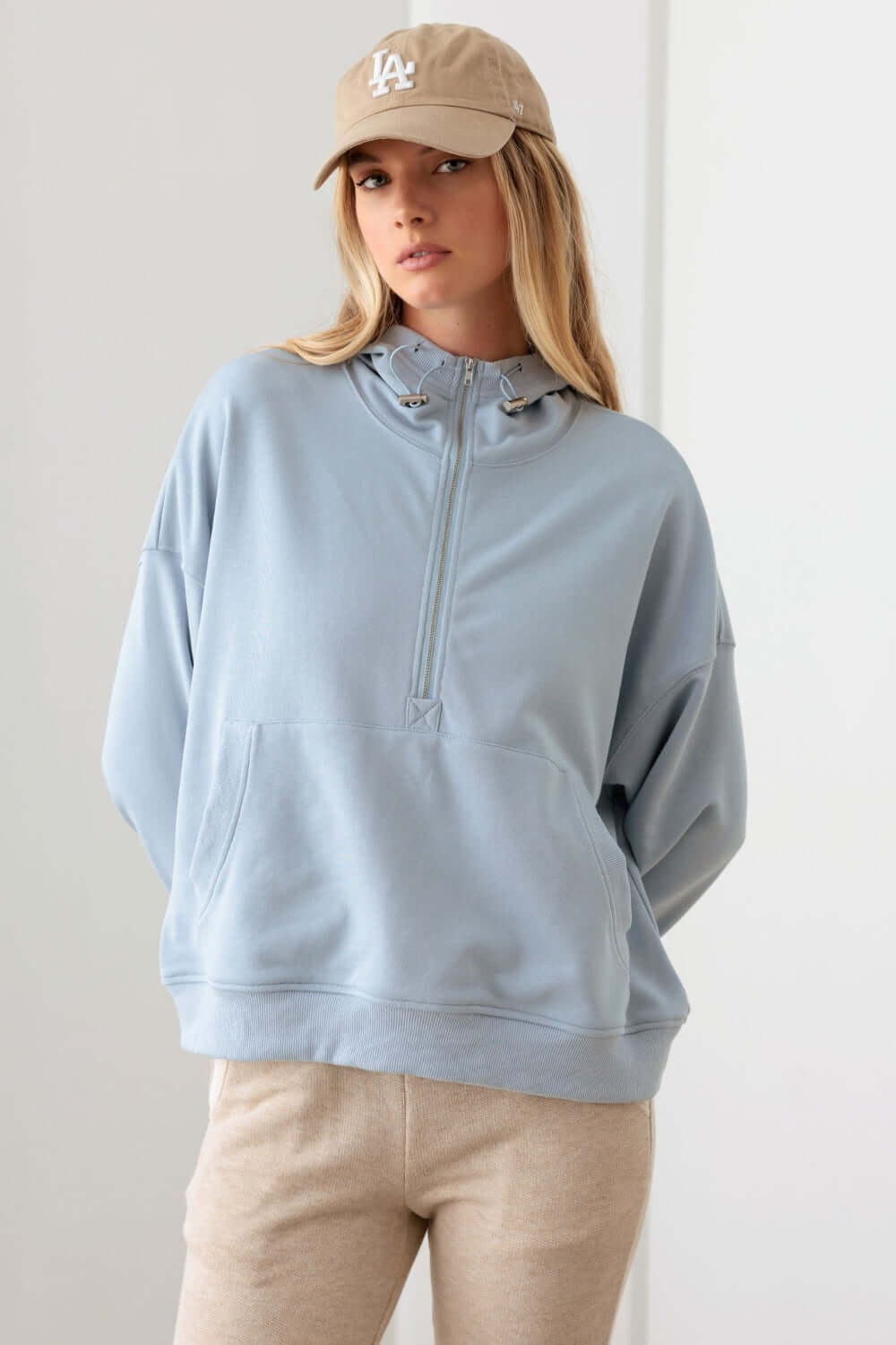 Woman wearing a light blue Half Zip Drawstring Mock Neck Hoodie with front pocket paired with beige pants and a beige cap.