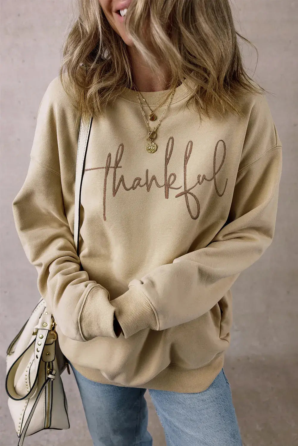 Woman wearing Bella Road Thankful Round Neck Long Sleeve Sweatshirt in beige, paired with blue jeans and a white handbag.