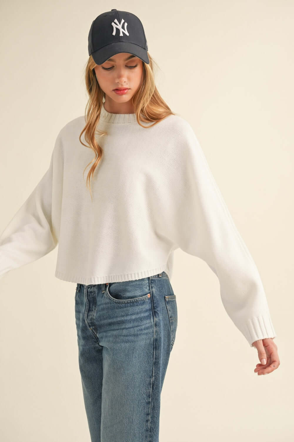 Woman wearing Bella Road Mable Round Neck Dolman Sleeve Cropped Sweater with blue jeans and black baseball cap