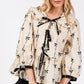 SAGE + FIG Bow Print Tie Front Babydoll Blouse in cream with black bows, featuring a tie-front and flowy silhouette.