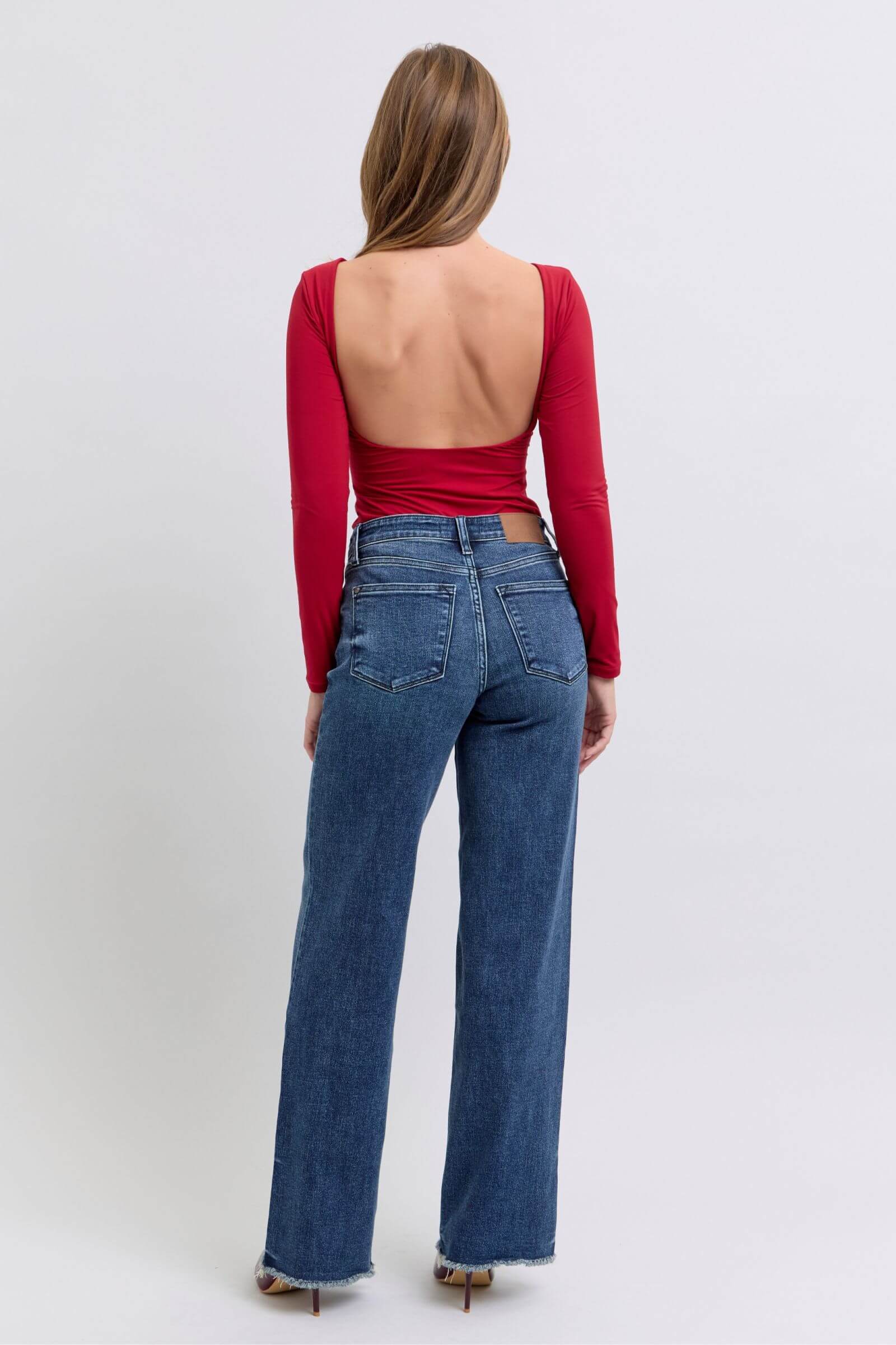 Woman in red backless top wearing Judy Blue raw hem mid-rise jeans, showcasing a trendy casual style.