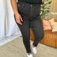 Woman wearing RFM Full Size High Rise Tummy Control Skinny Jeans in black, standing in light-filled room with sofa in background