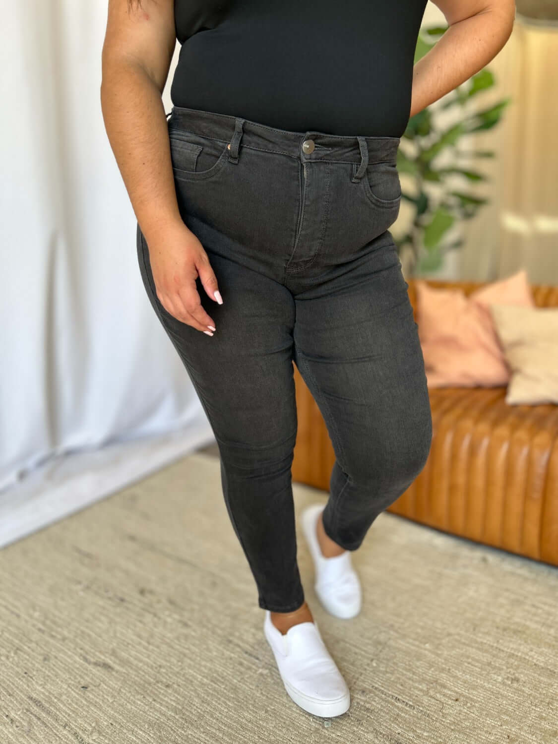 Woman wearing RFM Full Size High Rise Tummy Control Skinny Jeans in black, standing in light-filled room with sofa in background