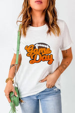 Woman wearing white GAME DAY round neck short sleeve T-shirt with orange lettering and carrying a green bag.