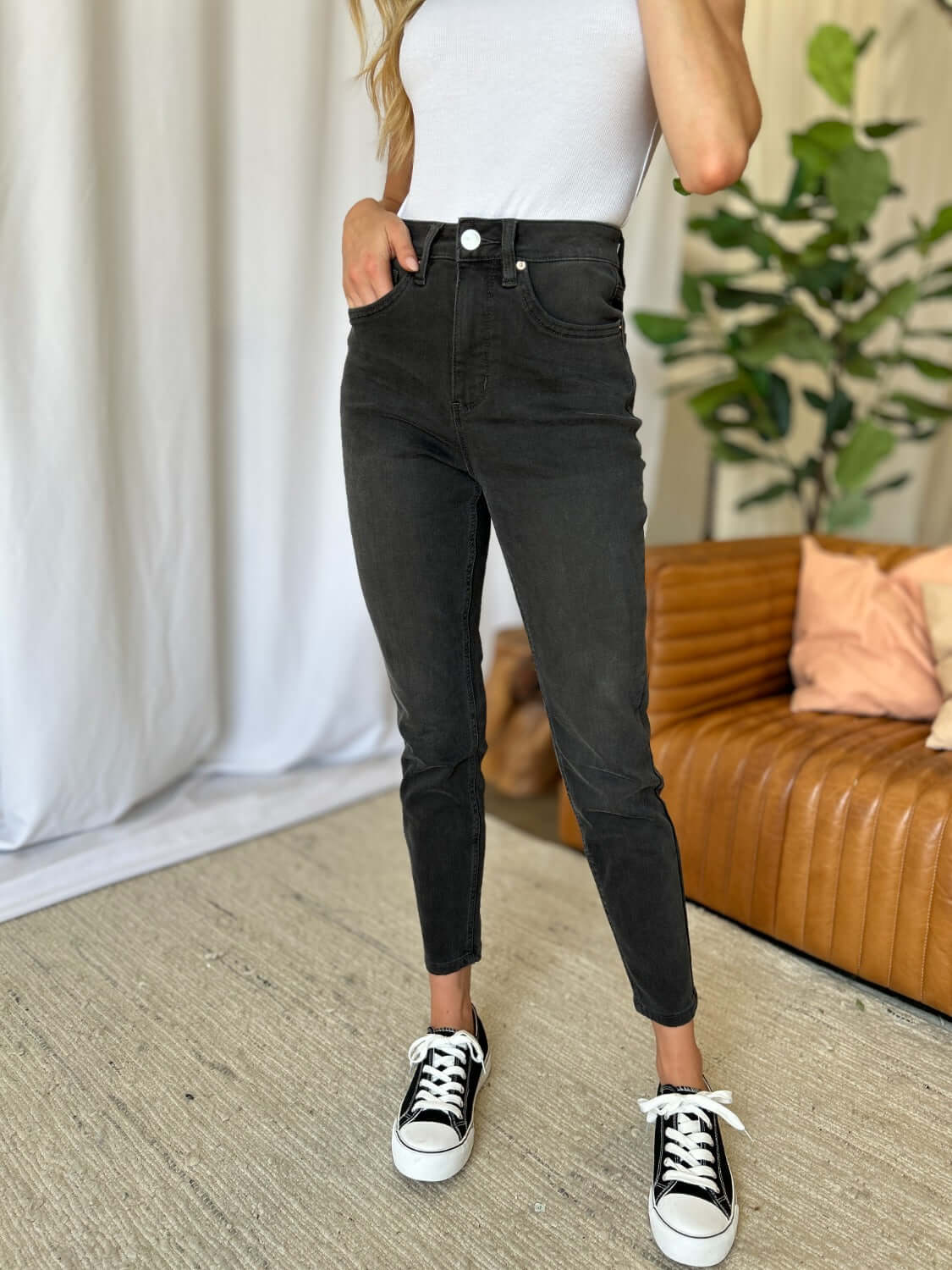 Woman wearing RFM Full Size High Rise Tummy Control Skinny Jeans with a white top and black sneakers in a stylish living room.