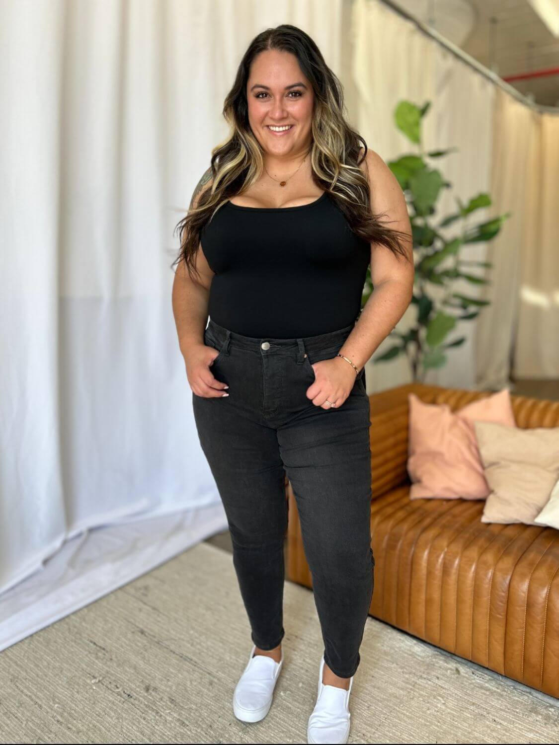 Woman wearing RFM Full Size High Rise Tummy Control Skinny Jeans with a black top, standing confidently in a cozy, modern room.