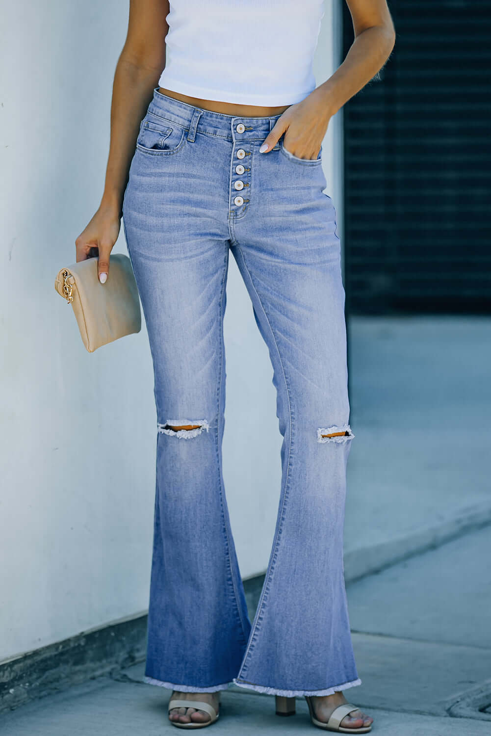 Woman wearing Bella Road Button-Fly Distressed Raw Hem Flare Jeans with a white top and holding a beige clutch.