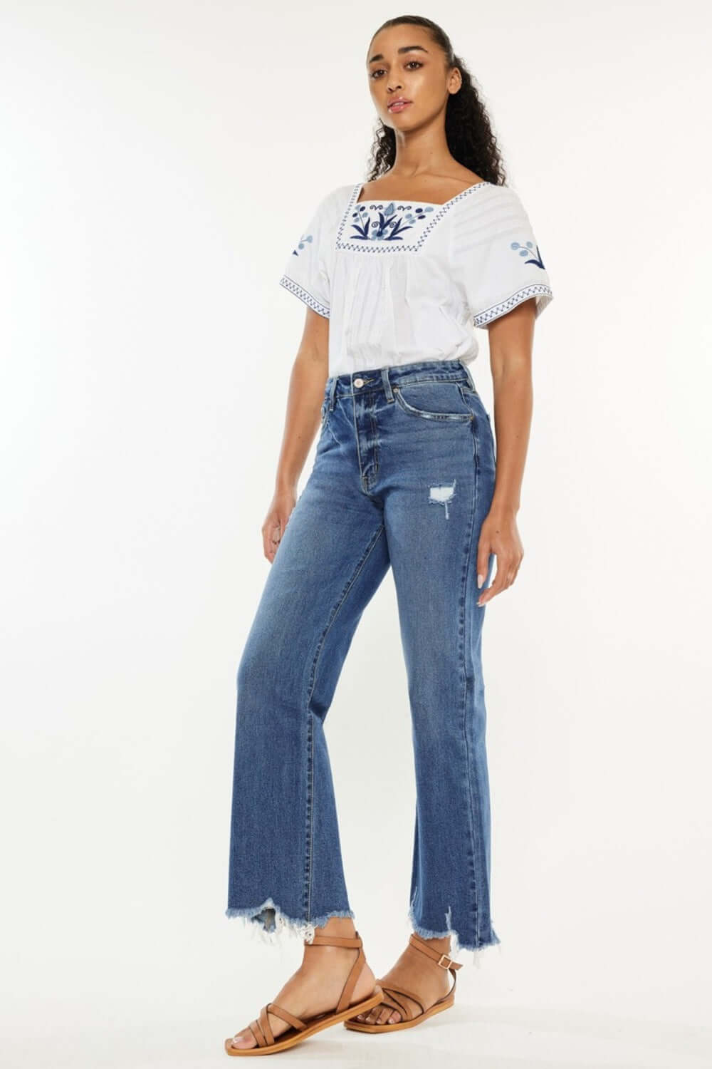 Woman wearing Kancan high-rise slim wide leg jeans in medium wash with trendy hem details, paired with a stylish blouse and sandals.