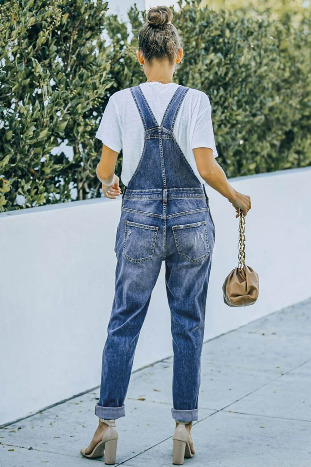 Woman wearing pocketed distressed denim overalls with acid wash outdoors, paired with a white t-shirt and beige high-heeled sandals.