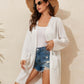 Woman wearing a white lace detail open front long sleeve cover-up over a white tank top, paired with denim shorts and a straw hat.