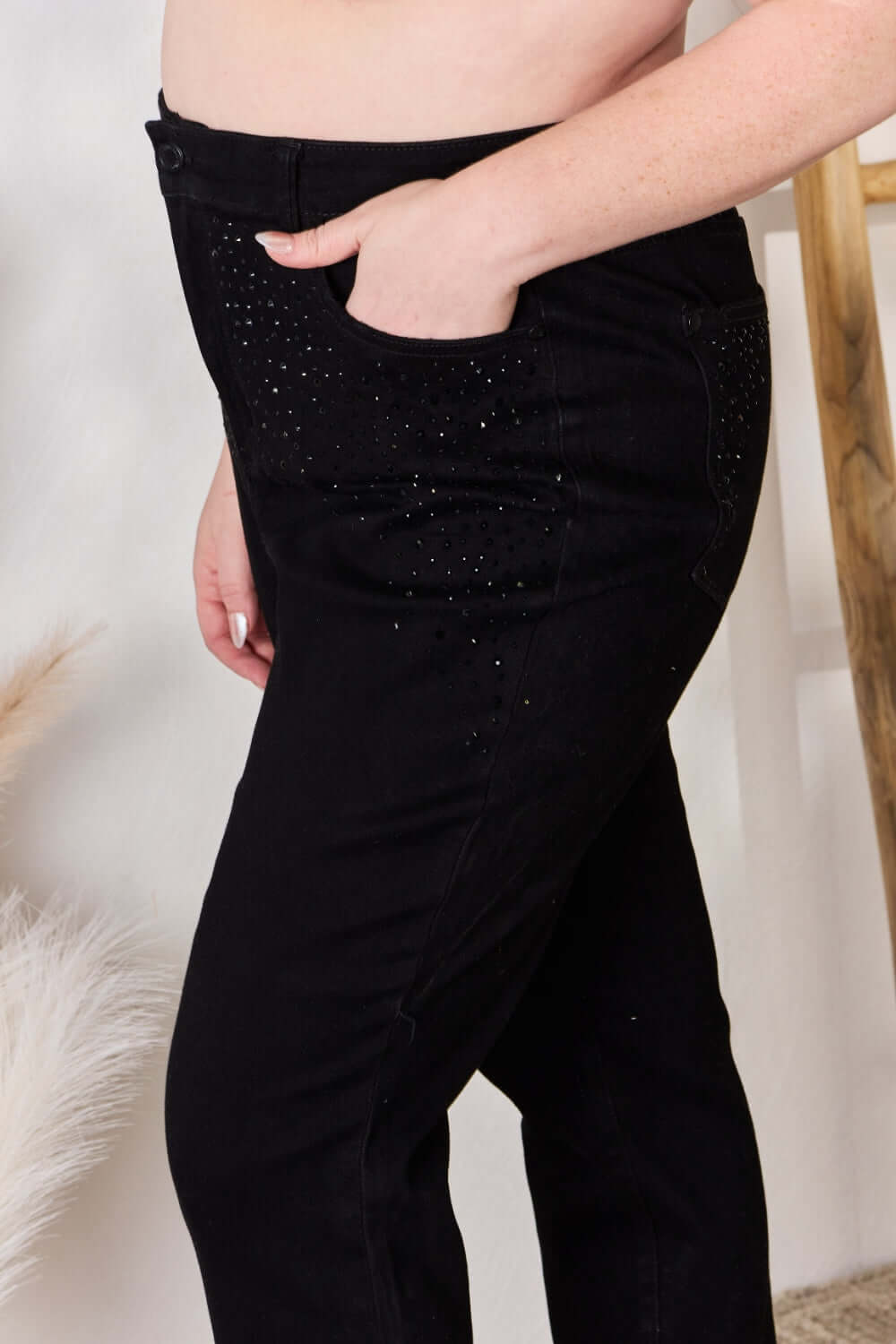 A person wearing black rhinestone embellished Judy Blue slim jeans with a high waist and hand in pocket.