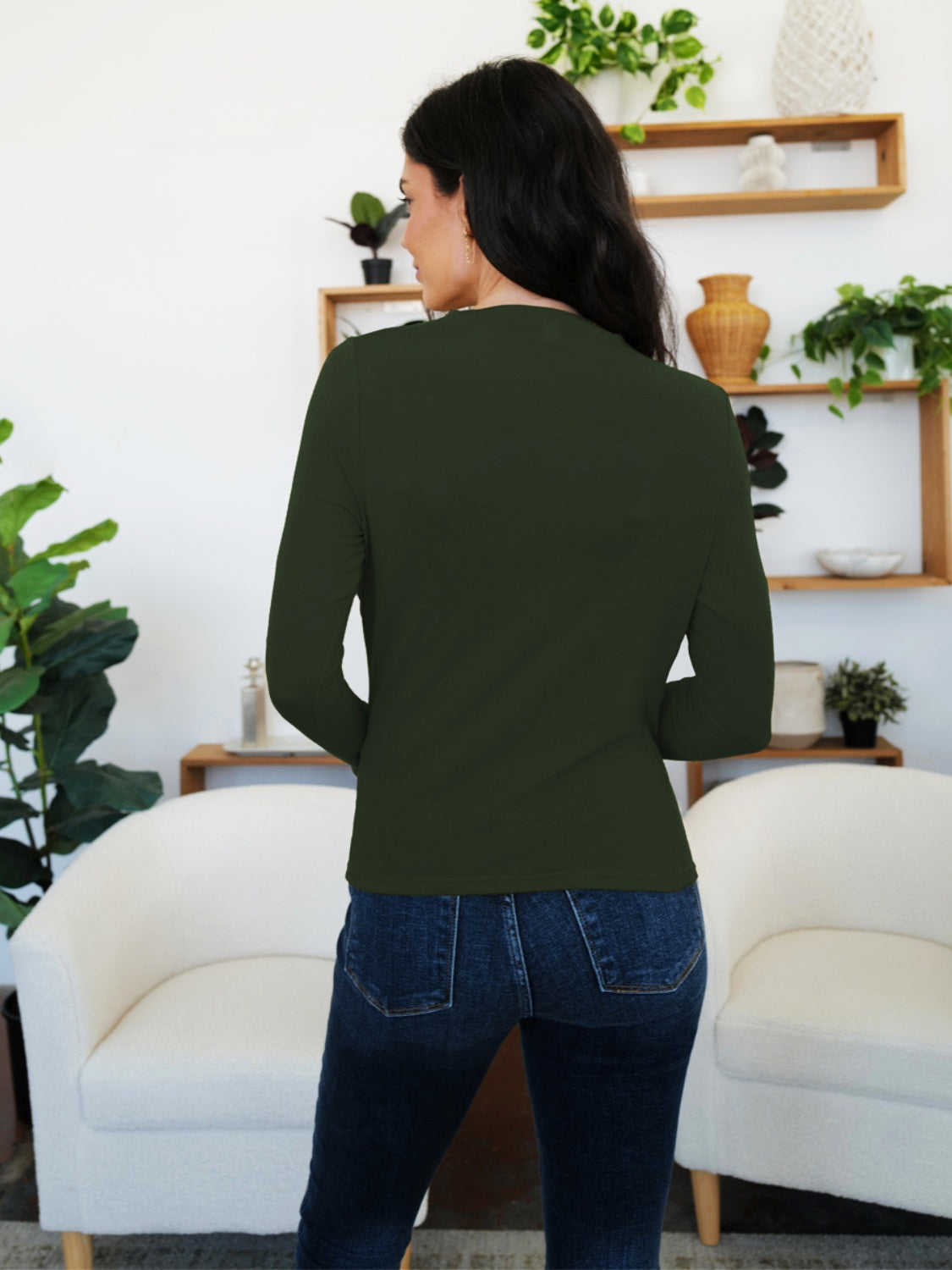 Woman wearing FAM-FAM Ruched Mock Neck Long Sleeve T-Shirt in Army Green, standing in stylish living room, showing back view.