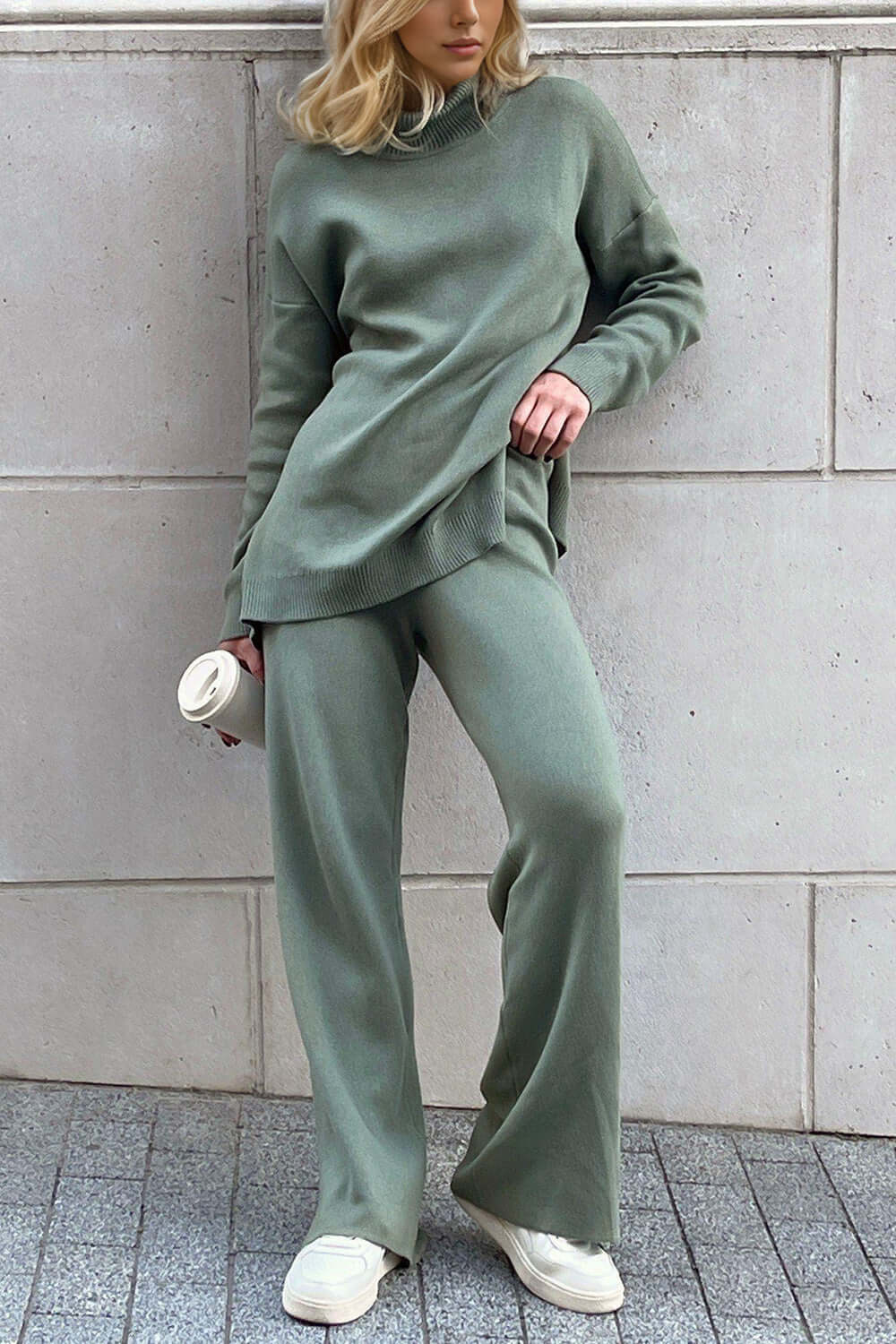 Stylish woman in green turtleneck side slit sweater and matching pants, holding coffee cup, showcasing cozy, relaxed fashion style.