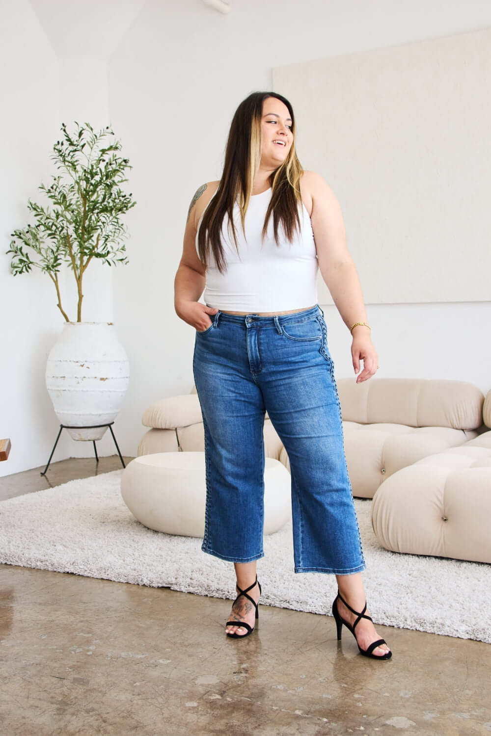 Woman wearing Judy Blue braid side detail wide leg jeans with a white top and black heels standing in a modern living room.