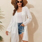 Woman wearing a white lace detail open front long sleeve cover-up over a white top and denim shorts, accessorized with a floppy hat and sunglasses.