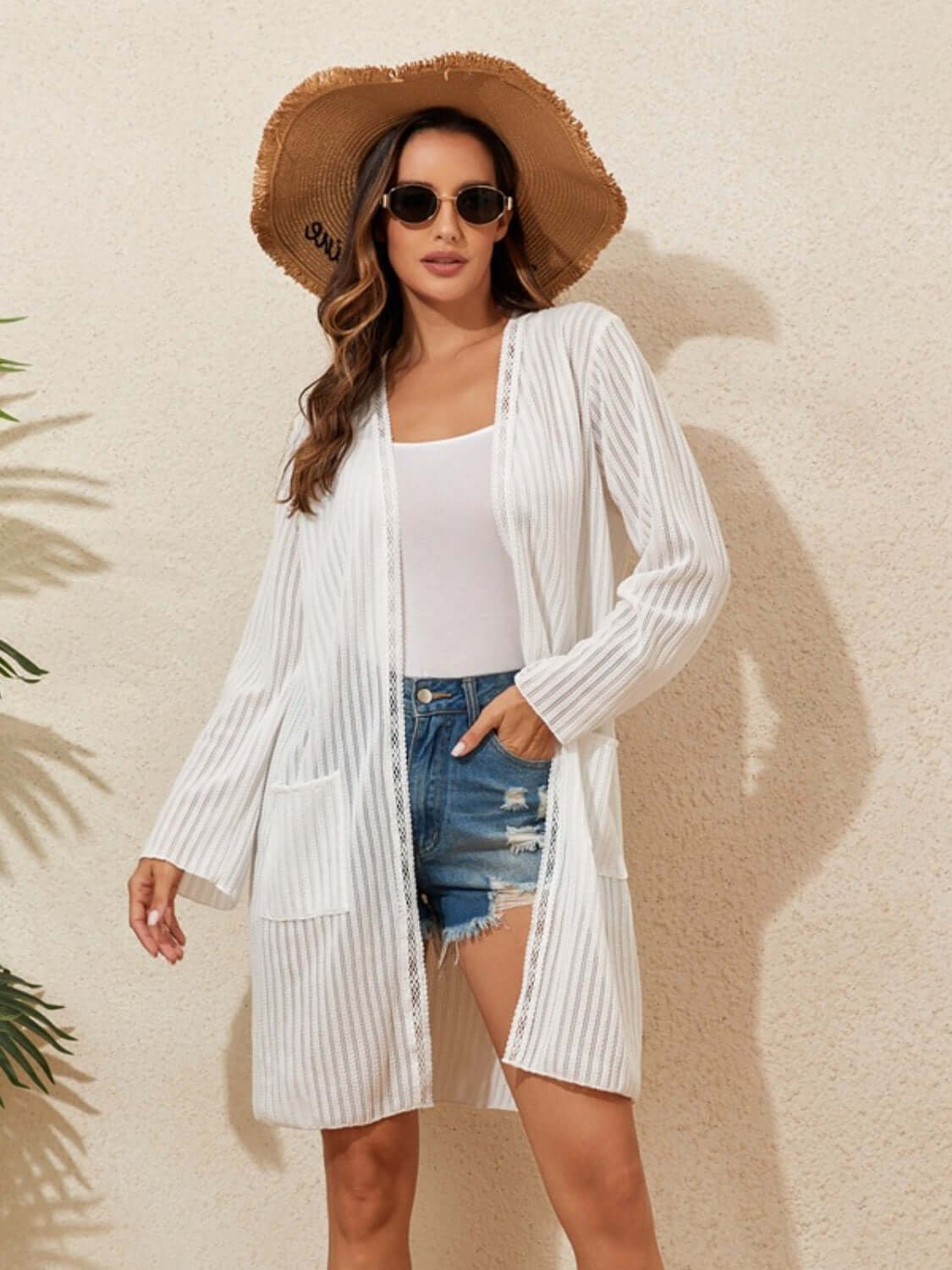 Woman wearing a white lace detail open front long sleeve cover-up over a white top and denim shorts, accessorized with a floppy hat and sunglasses.