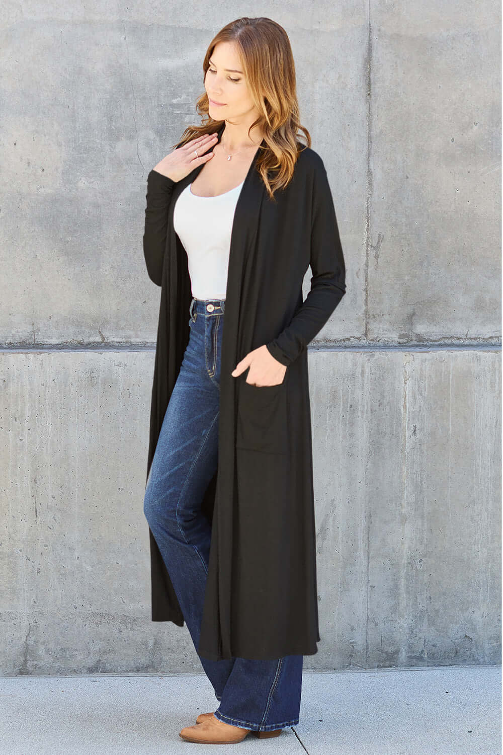 Woman wearing black open front long sleeve cover up with pockets over white top and blue jeans, standing against concrete wall.