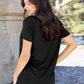 Woman wearing Basic Bae Bamboo full size black V-neck short sleeve T-shirt, standing outdoors with a serene backdrop.