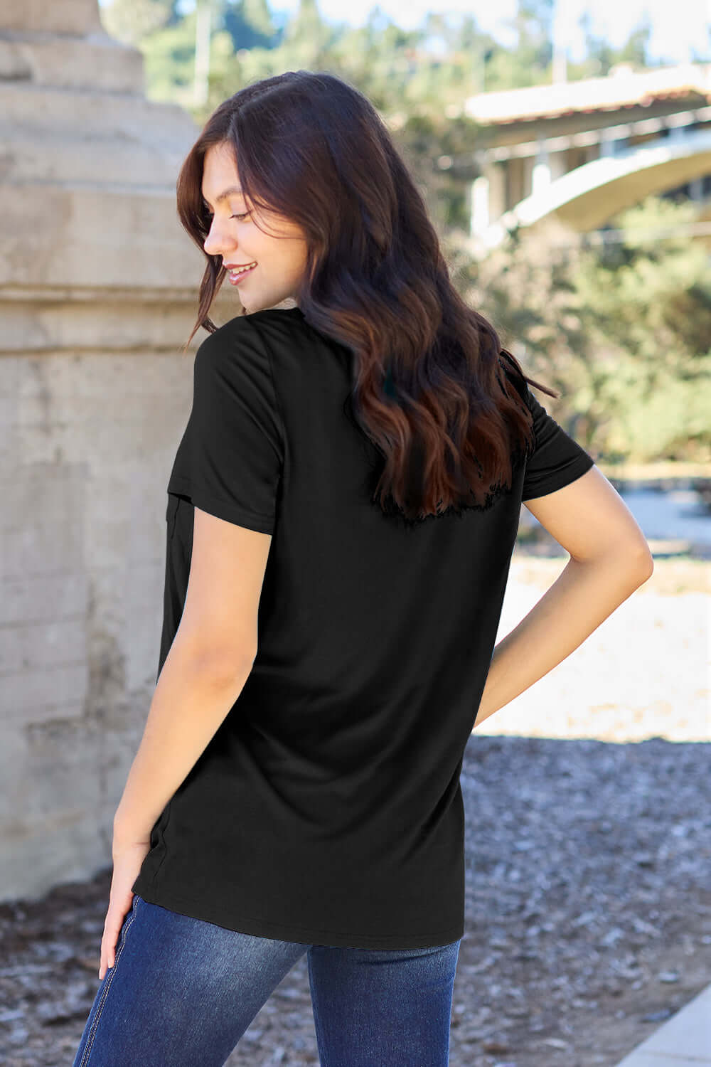 Woman wearing Basic Bae Bamboo full size black V-neck short sleeve T-shirt, standing outdoors with a serene backdrop.