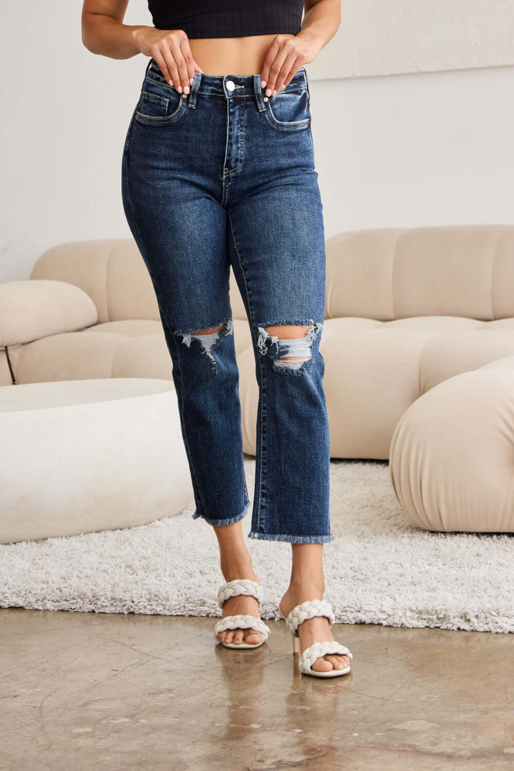 Woman wearing RFM Jeans Tummy Control Distressed High Waist Raw Hem Jeans standing in modern living room.