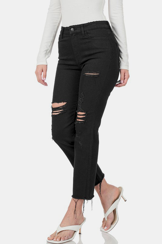 Black distressed cropped jeans with raw hem, styled casually with white heels and a long-sleeve top, perfect for edgy fashion looks.