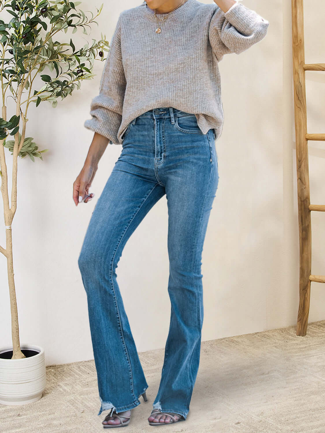 Woman wearing distressed raw hem bootcut jeans with pockets and a grey sweater standing indoors.