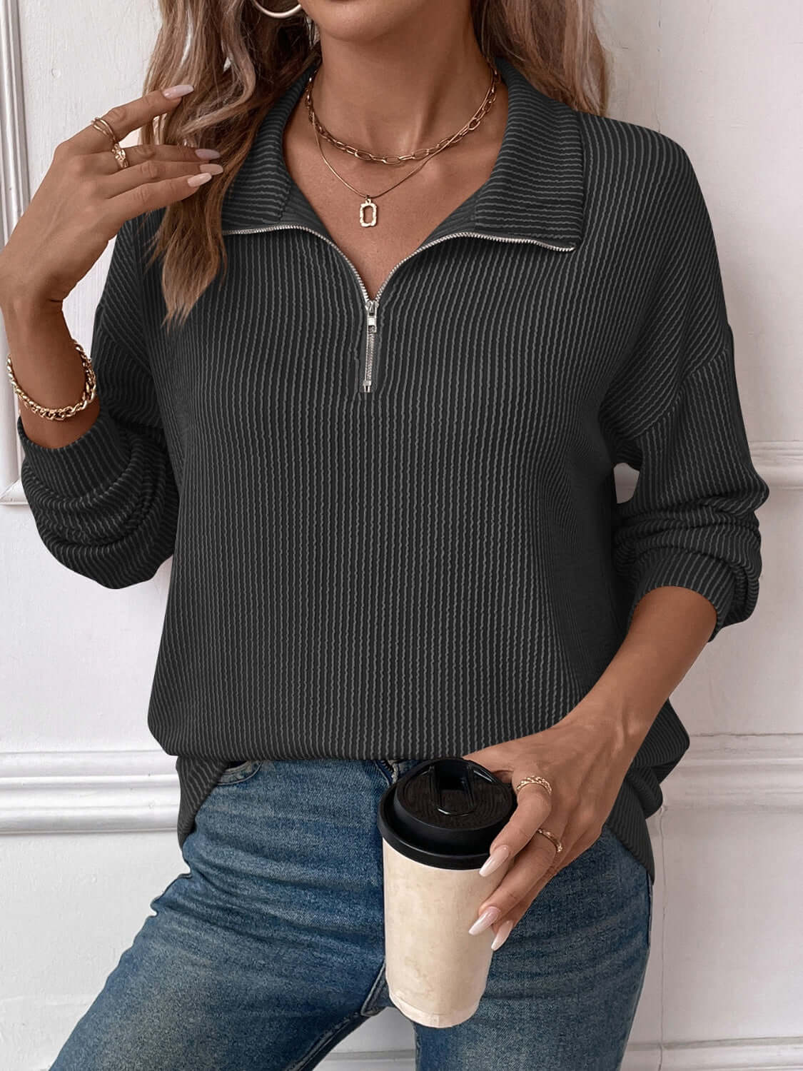 Woman wearing a striped half zip long sleeve t-shirt in dark gray with blue jeans, holding a coffee cup, and accessorized with gold jewelry.